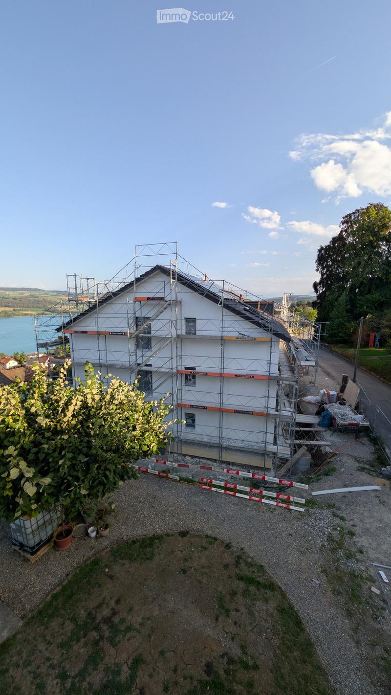 A two-story house under construction with scaffolding, located near a body of water, surrounded by greenery and construction materials.