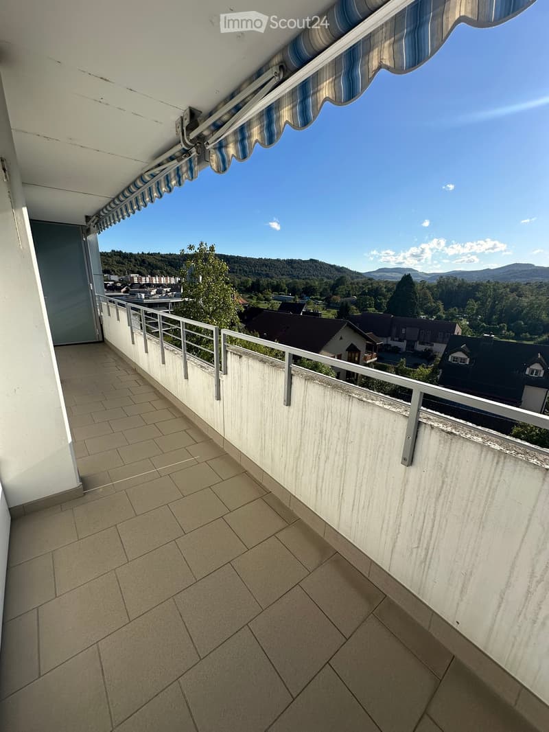 Balcony with tiled floor, metal railing, and striped awning, overlooking a scenic landscape with houses and hills.