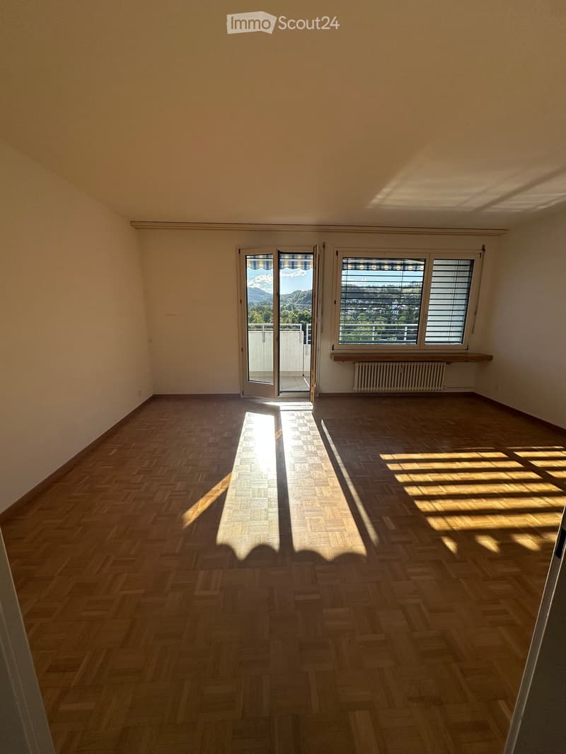 Empty room with wooden herringbone flooring, large windows, and a sliding glass door leading to a balcony.