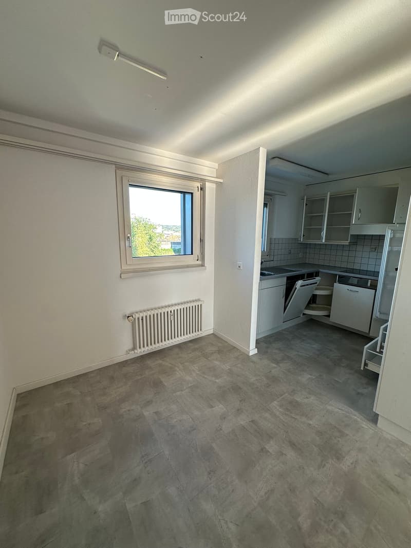 Empty room with tiled floor, white walls, radiator, window, and adjacent kitchen area with cabinets and appliances.