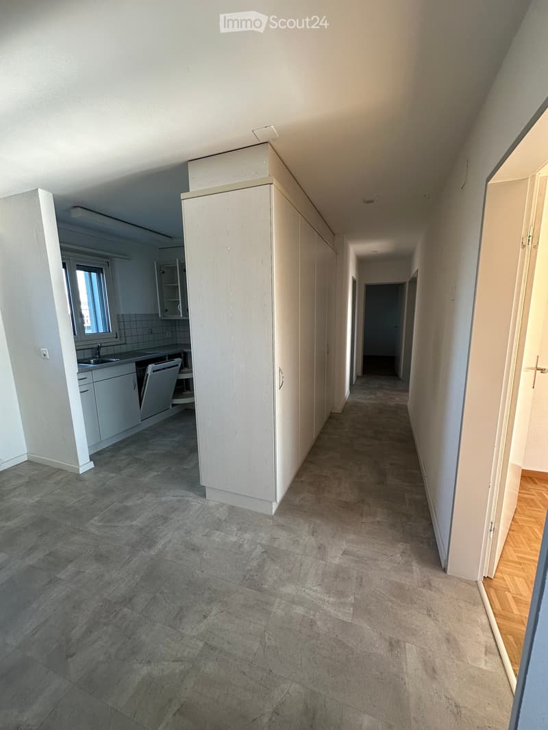 Empty hallway with tiled floor, white walls, and doors leading to other rooms. A kitchen area is visible to the left with cabinets, sink, and dishwasher.