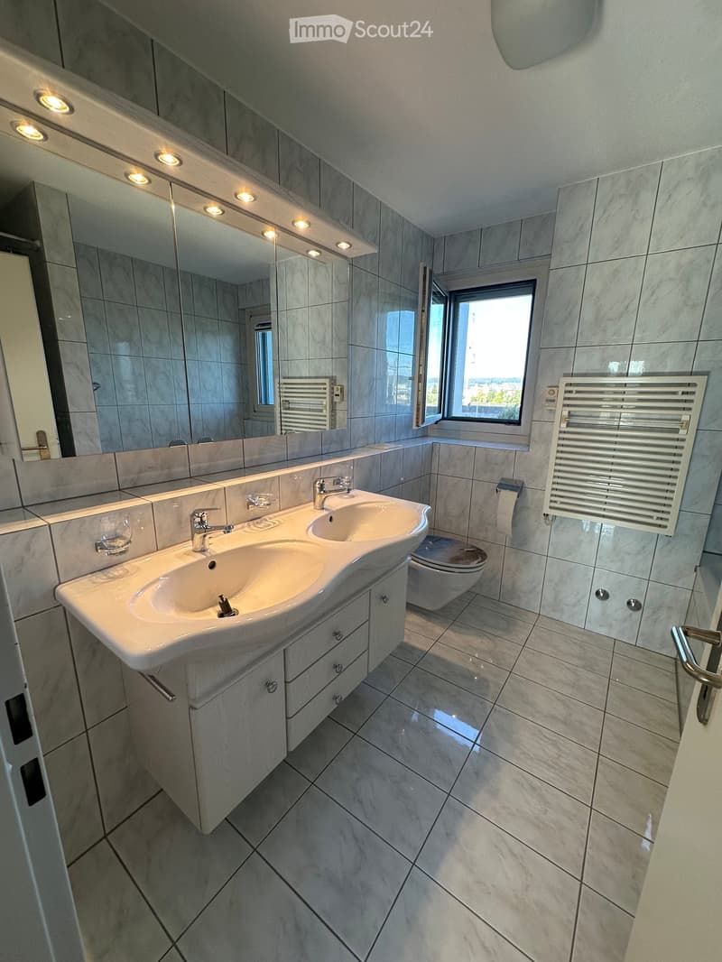 Modern bathroom with double sink, large mirror, toilet, radiator, and a window letting in natural light.