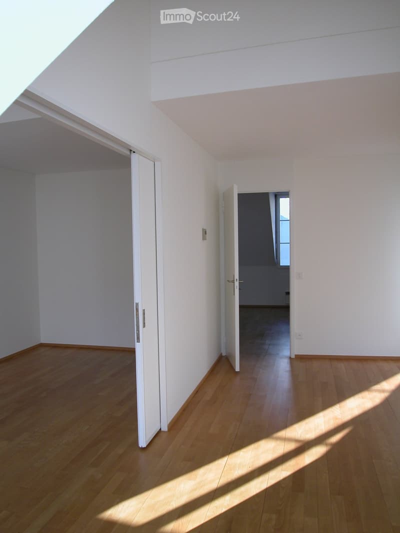 Empty room with light wooden flooring and white walls, featuring two open doors leading to adjacent spaces.