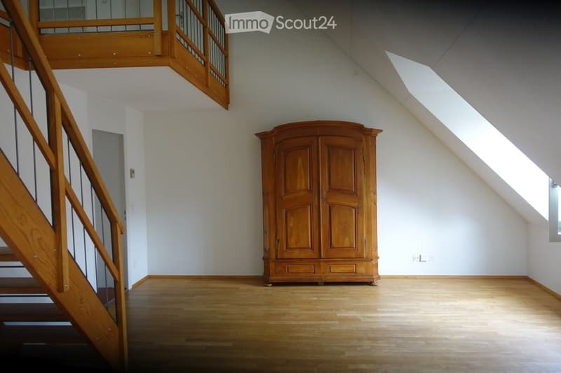 Spacious attic room with wooden flooring, white walls, and a large wooden wardrobe. A wooden staircase leads to an upper level with a wooden railing.