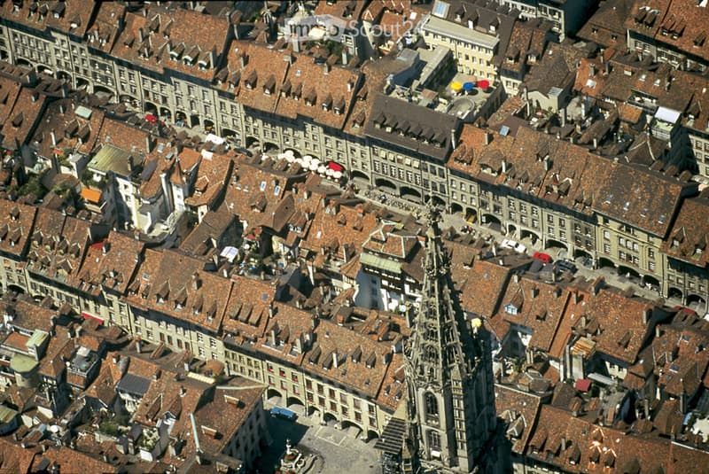 Aerial view of a historic European city with numerous buildings featuring red-tiled roofs, narrow streets, and a prominent church spire in the center.
