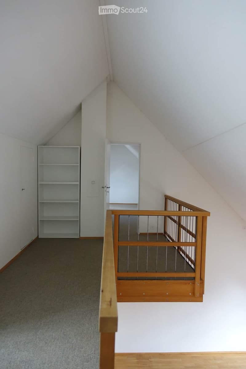 Empty hallway with wooden flooring, white walls, and a wooden railing leading to an open area. A white shelving unit is visible on the left side.