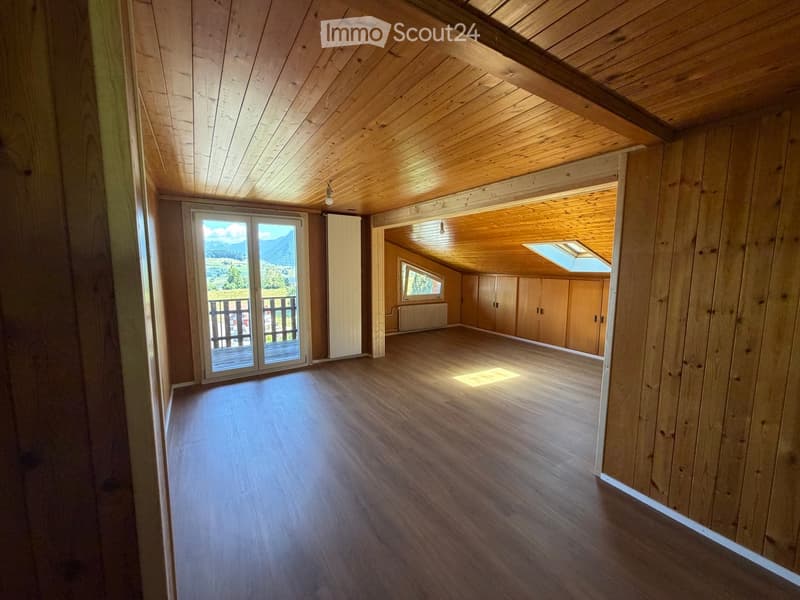 Empty room with wooden walls, wooden floor, exposed wooden ceiling beams, large window with a view, and built-in wooden cabinets.