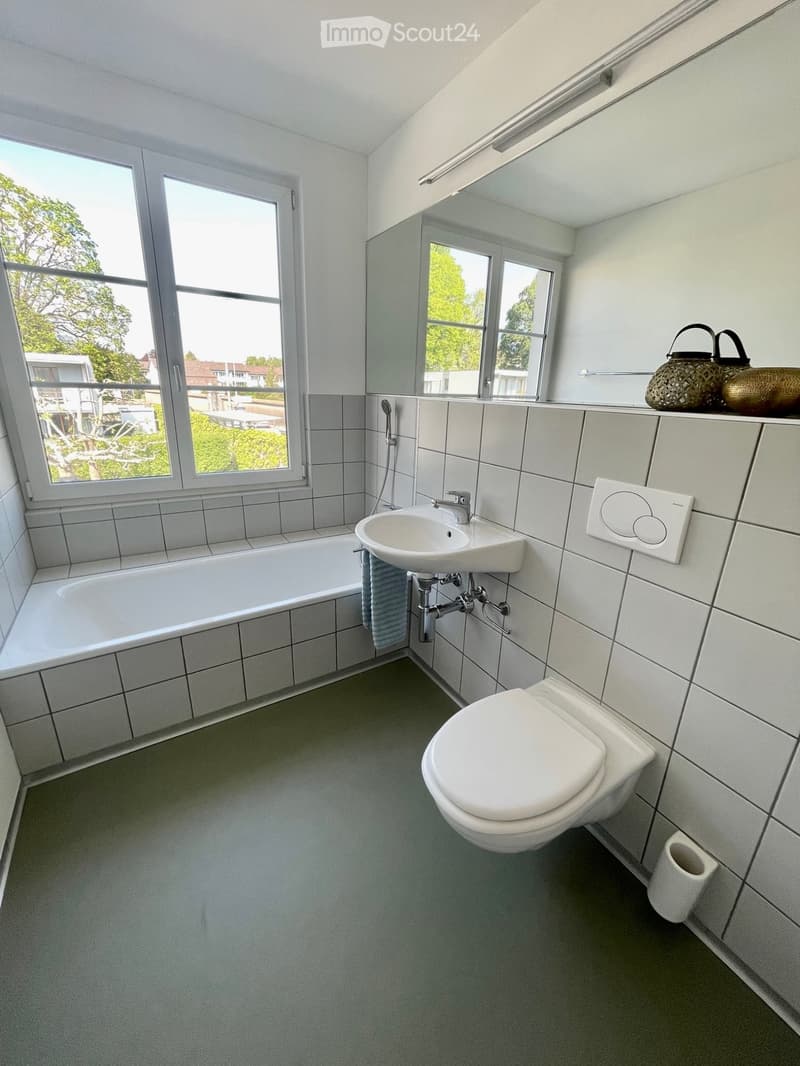 Modern bathroom with white tiles, bathtub, sink, toilet, and decorative baskets on the mirror shelf. Large windows provide natural light.