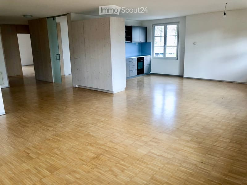Empty room with wooden flooring, white walls, and a small kitchen area visible in the background.