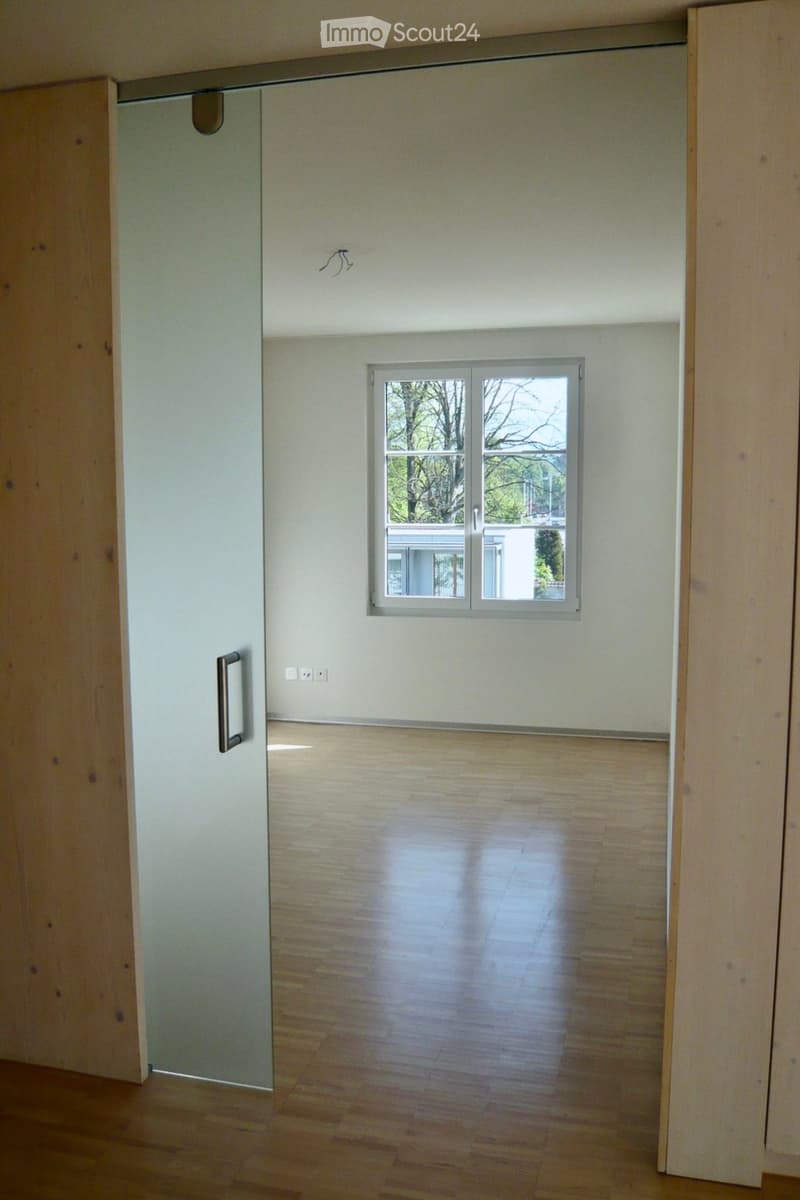 Empty room with wooden flooring, white walls, and a window with a view of trees and a neighboring building.