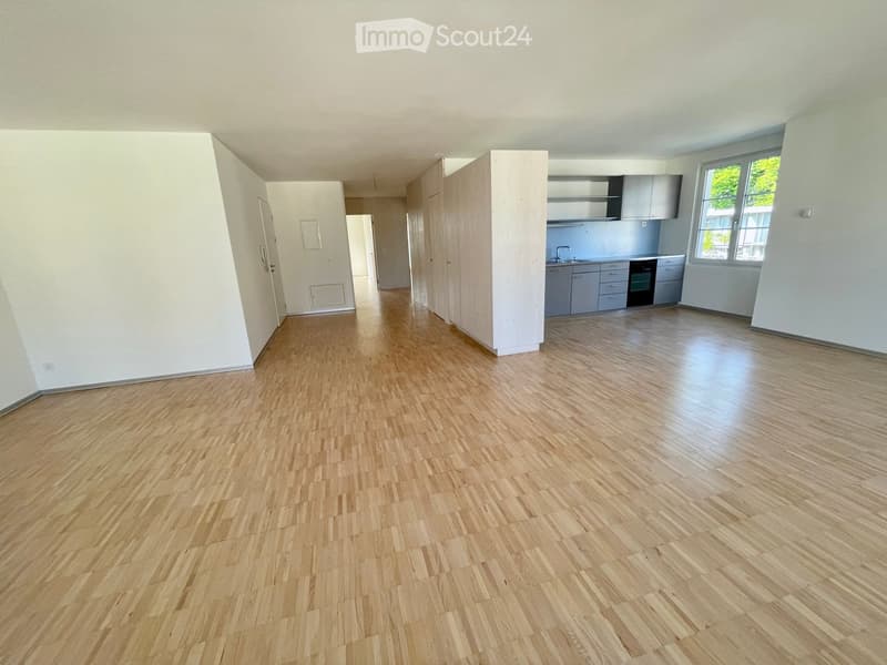 Empty room with light wooden flooring, white walls, and a small kitchen area with cabinets and appliances visible in the background.