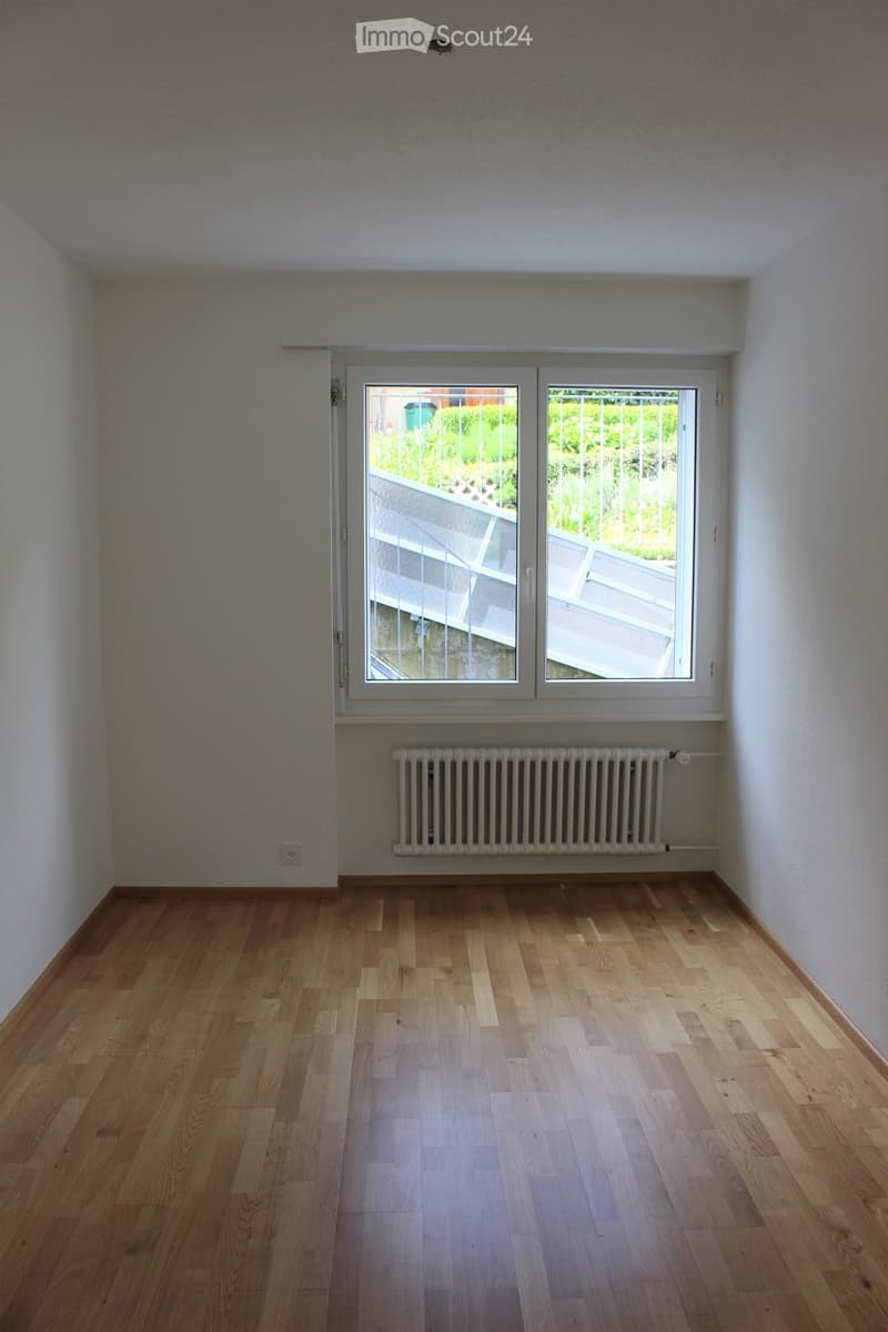 Empty room with wooden flooring, white walls, large window with view of outdoor area, radiator below window, and visible electrical outlet on wall.
