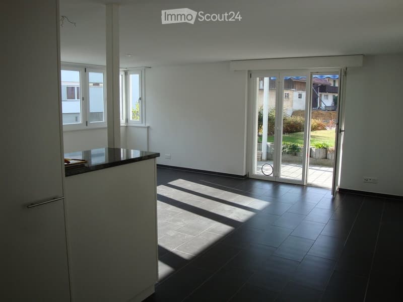 Empty room with dark tiled floor, white walls, large windows, and sliding glass doors leading to a garden area.