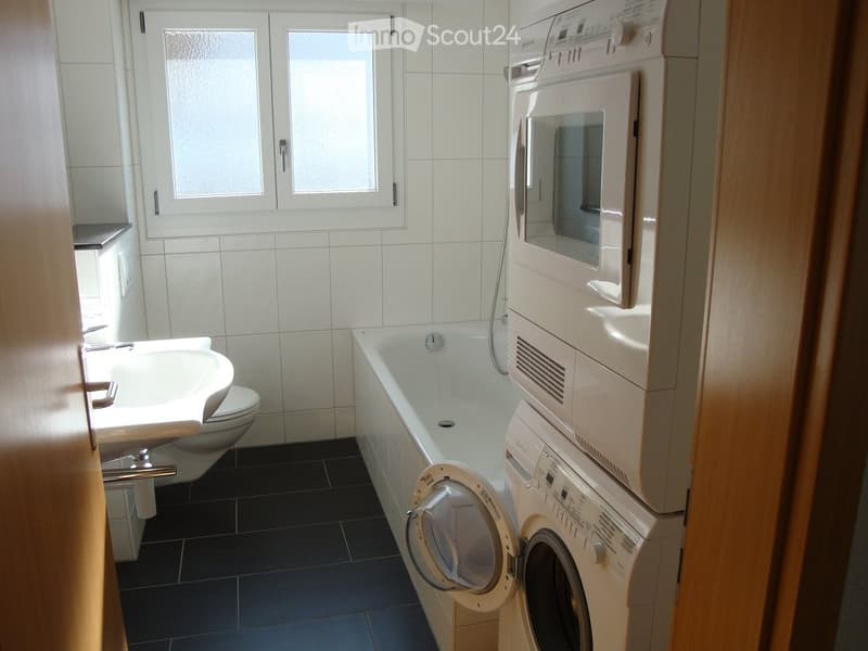 Modern bathroom with white tiles, a bathtub, sink, toilet, and stacked washing machine and dishwasher.