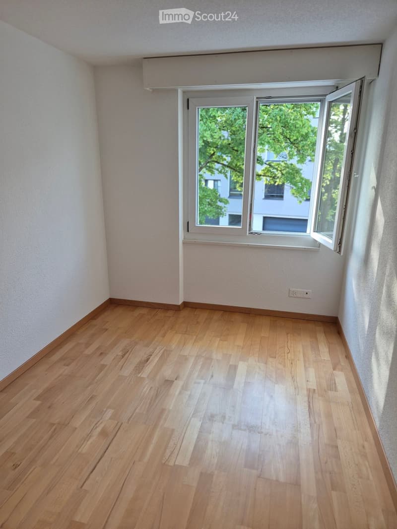 Empty room with wooden flooring, white walls, and a large window with white frame and blinds partially open, showing greenery outside.