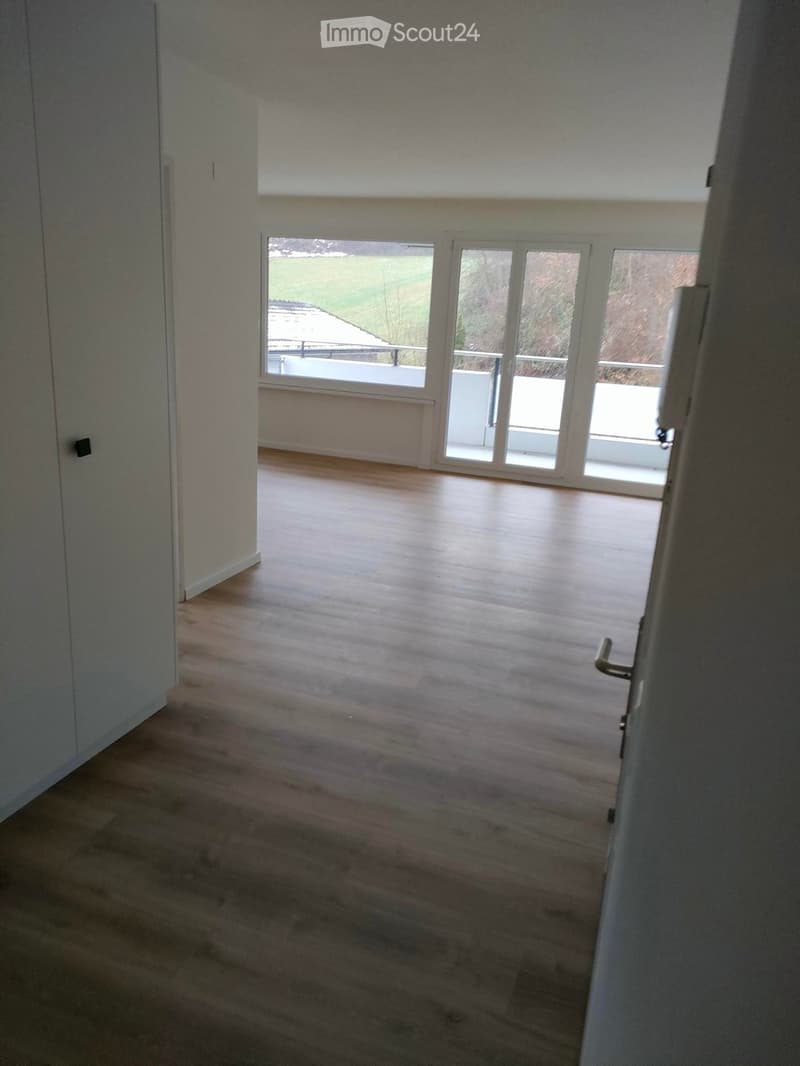 Empty room with wooden flooring, white walls, and large windows leading to a balcony area.