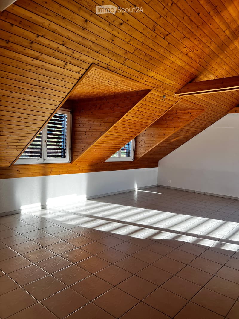 Empty room with wooden slanted ceiling, tiled floor, and two windows with blinds allowing natural light to enter.