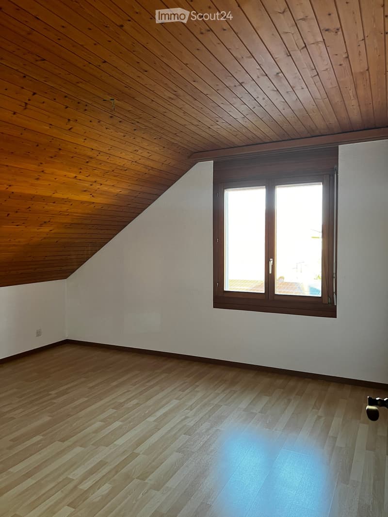 Empty room with wooden flooring and slanted wooden ceiling, featuring a large window with brown frame allowing natural light to enter.