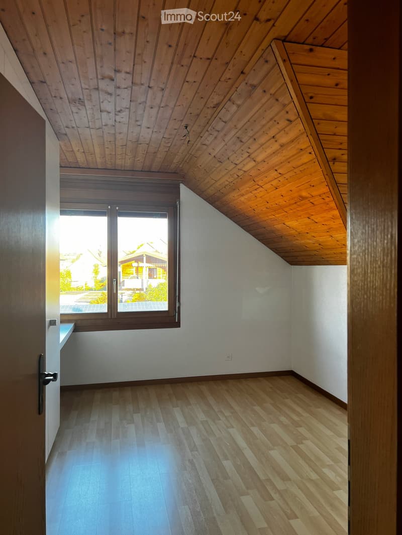 Empty attic room with wooden slanted ceilings, wooden flooring, and a large window offering a view of the outdoor area.