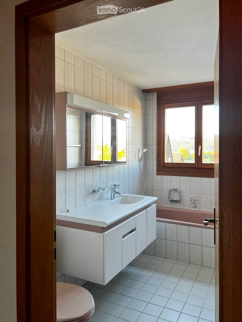Modern bathroom with white tiles, a white sink with a chrome faucet, a mirrored cabinet, a bathtub, and a toilet. The room has large windows offering a scenic view.