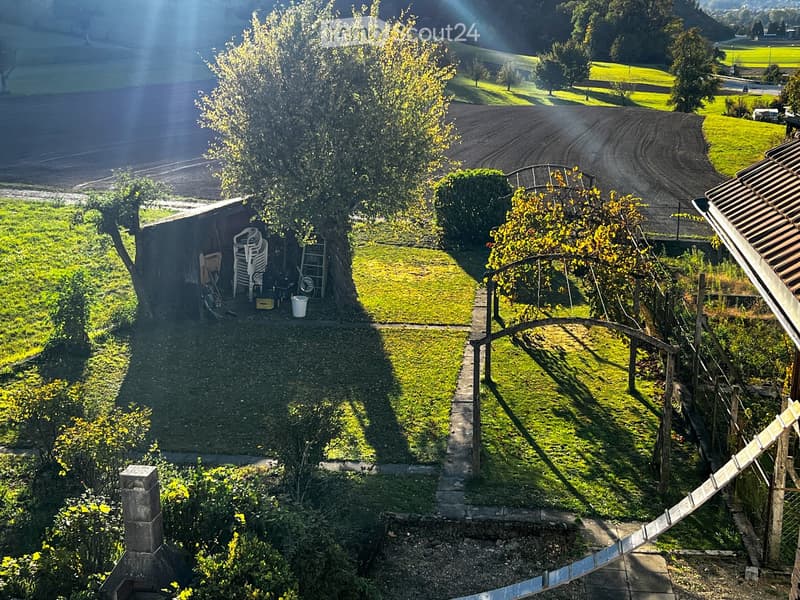 A well-maintained garden with a grassy area, a small shed, and a pathway leading to a wooden structure. The area is surrounded by trees and has a peaceful, rural setting with visible farmland in the background.