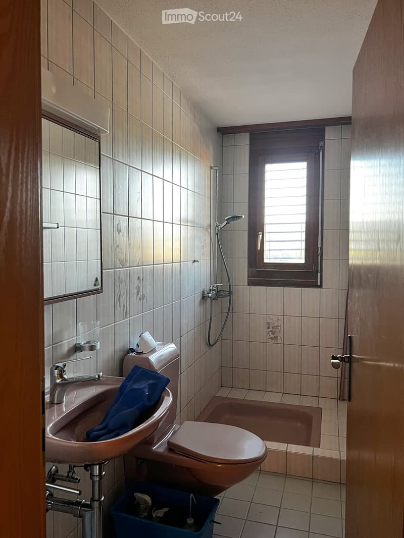 Bathroom with beige tiles, a sink with a blue towel, a toilet, a bathtub, a showerhead, a mirror, and a window with blinds.