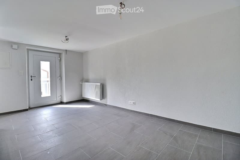 Empty room with tiled floor, white walls, a door with a window, a radiator, and light fixtures on the ceiling.