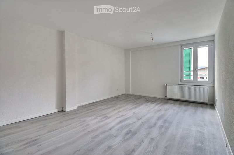 Empty room with light-colored wooden flooring, white walls, and a single window with green shutters. A radiator is visible below the window.