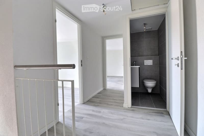 A modern hallway with light wooden flooring, white walls, and a staircase leading upwards. A small bathroom is visible to the right, featuring a toilet, sink, and dark tiled walls.