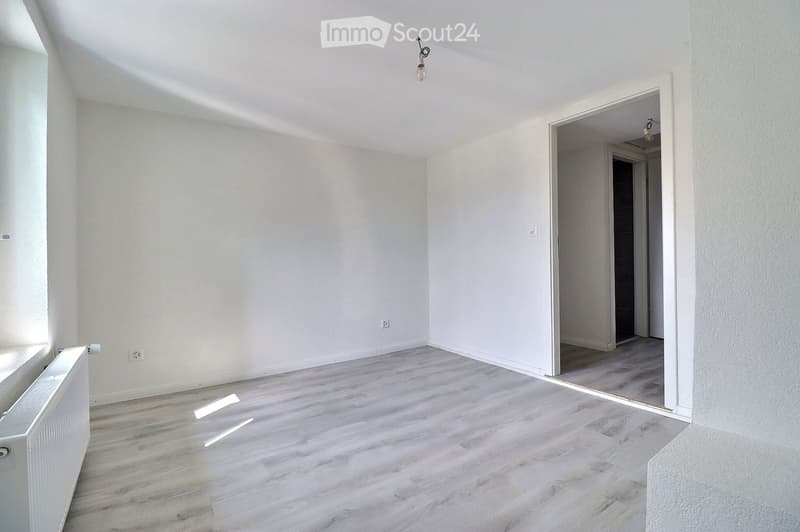 Empty room with light wooden flooring, white walls, and a radiator on the left side. A doorway leads to another room.