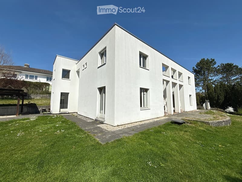 Modern two-story white house with large windows and a flat roof, surrounded by a well-maintained lawn and trees.