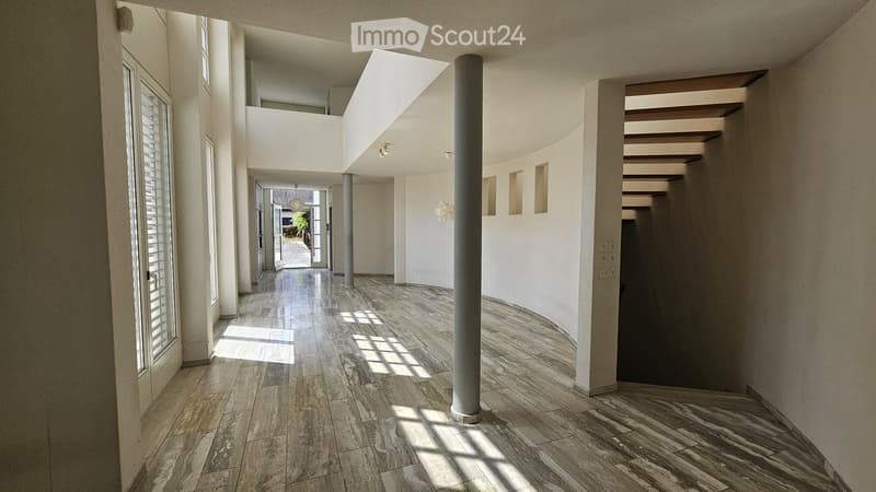 Bright, modern hallway with polished tile flooring, white walls, and large windows allowing natural light. Features a central column and a staircase leading upwards.