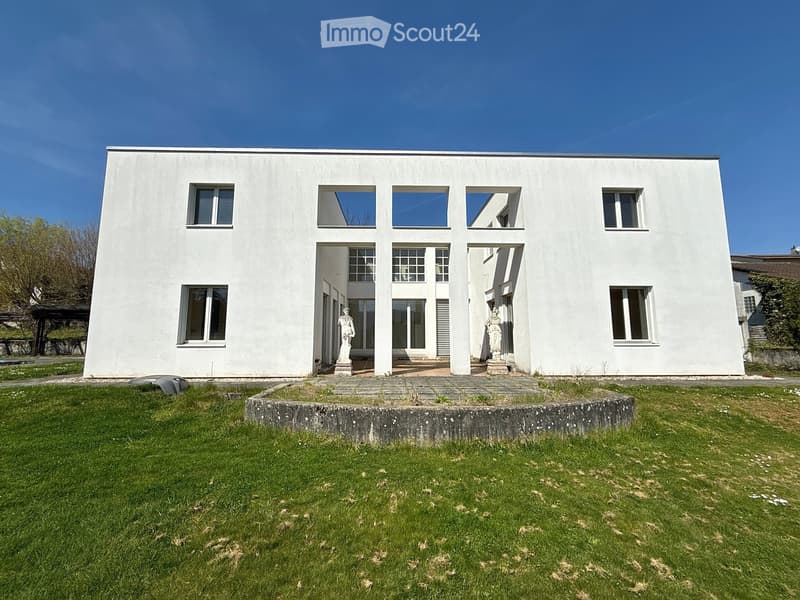 Modern white building with large windows and a flat roof, surrounded by a grassy lawn and statues at the entrance.