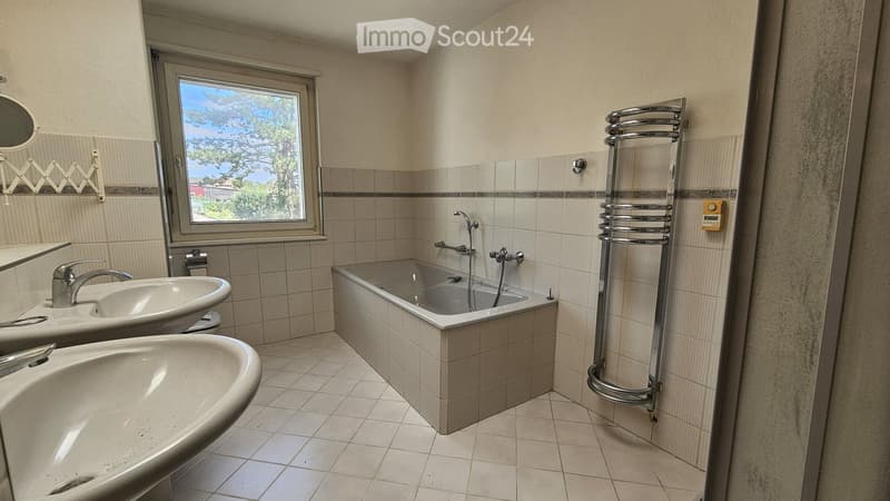 Modern bathroom with a bathtub, two sinks, a radiator, and a window offering a view of trees and a house outside.