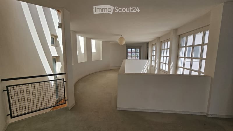 Empty hallway with beige walls, carpeted floor, large windows, and a hanging light fixture. The hallway features a curved design with a railing on one side and a white partition wall on the other.