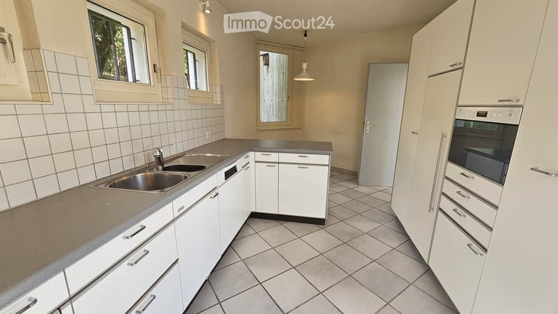 Modern kitchen with white cabinets, tiled walls, and a stainless steel sink. Features include an oven, a light fixture, and multiple windows allowing natural light.