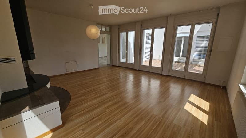 Bright, empty room with wooden flooring, large glass doors leading to a patio, and a modern fireplace on the left side.