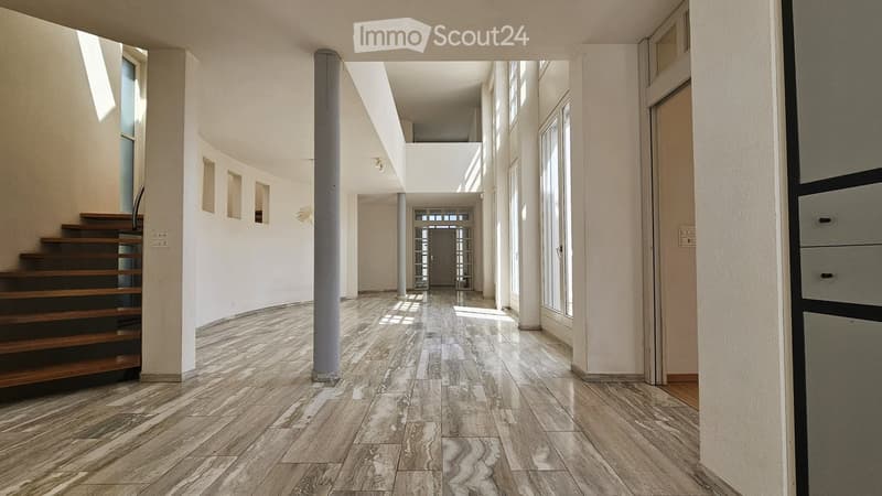Bright, modern hallway with polished marble flooring, white walls, and large windows allowing natural light. Features a wooden staircase on the left and a central column.