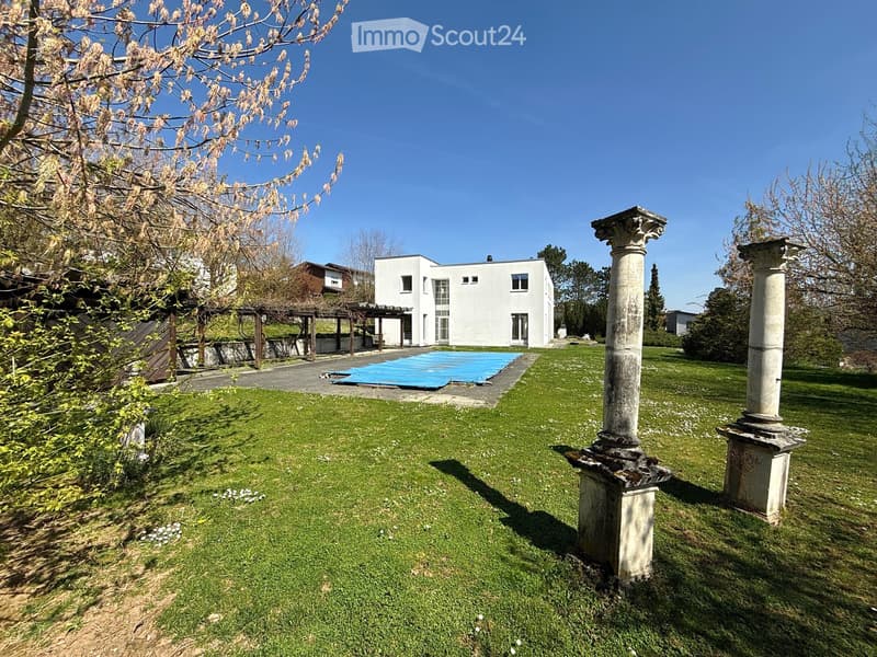 Well-maintained lawn with two stone columns, a covered patio area, and a modern white house in the background.