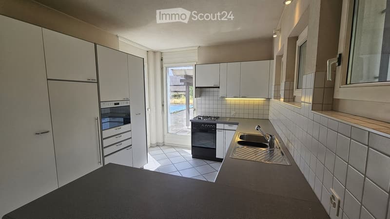 Modern kitchen with white cabinets, stainless steel sink, and appliances, featuring a view of the outdoor area through a large window.