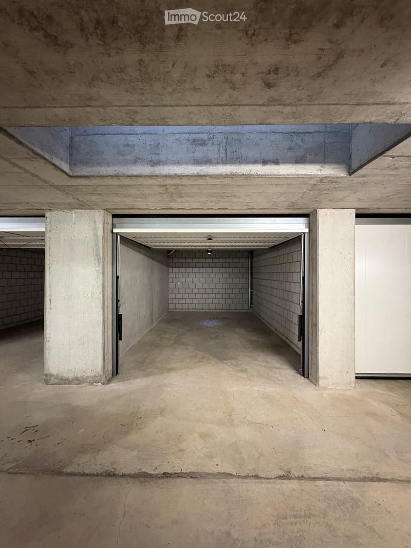 Empty underground garage with concrete walls, floor, and ceiling. Features a large open space with a single garage door at the far end.