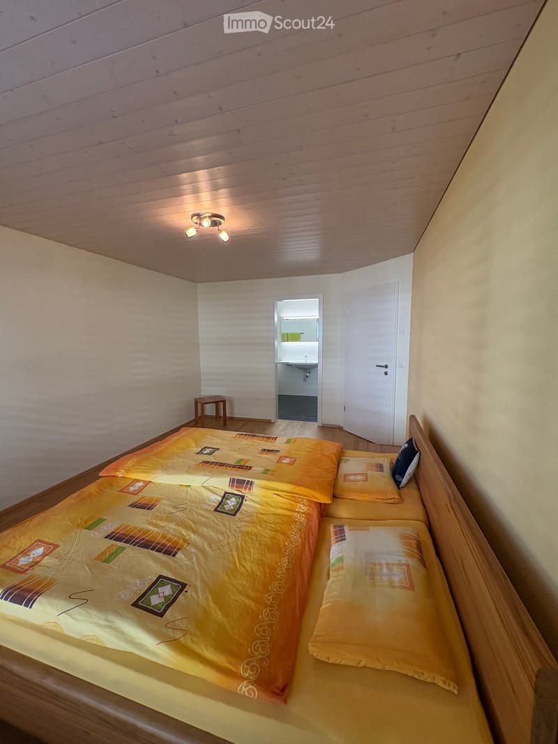 Cozy bedroom with a double bed, colorful bedding, a small wooden stool, and an adjacent bathroom visible through an open door.