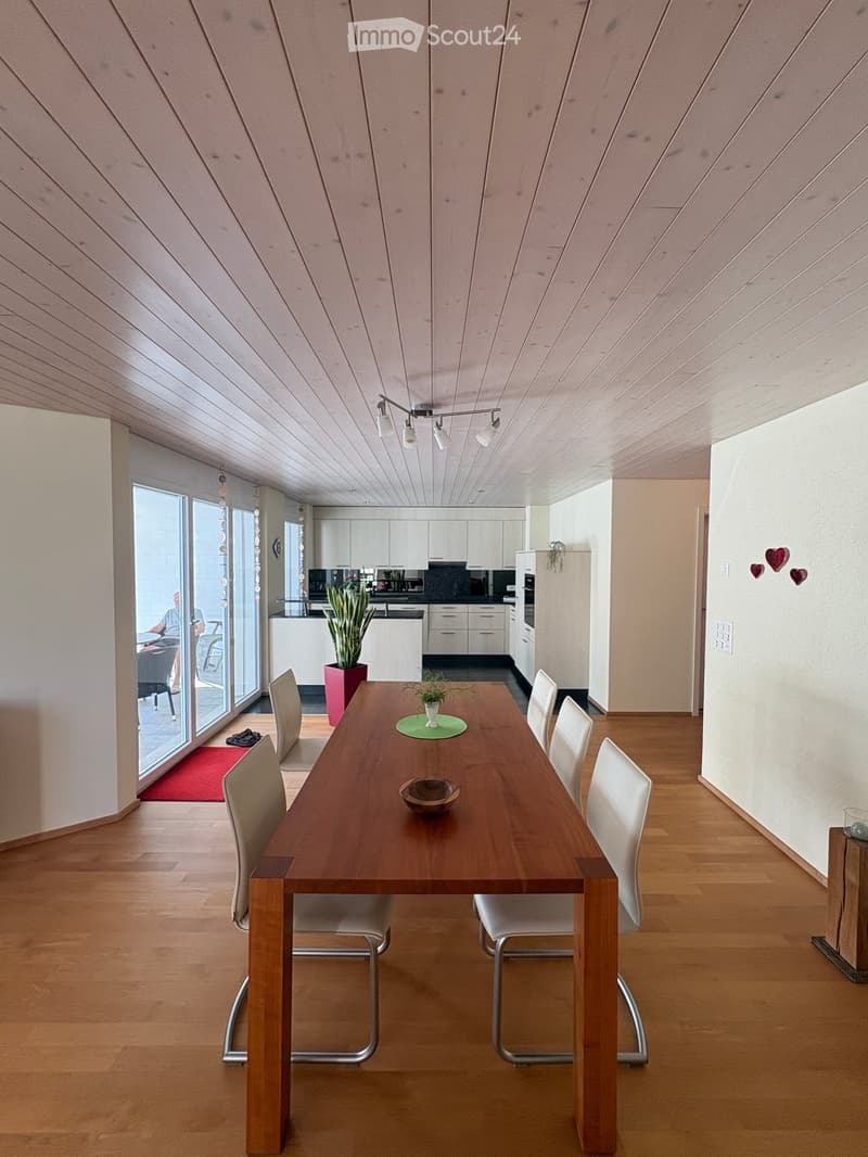 Modern dining room with wooden floor, slanted wooden ceiling, pendant lights, and a large wooden dining table surrounded by white chairs. A small plant and bowl are placed on the table. The room has large windows and an open kitchen area visible in the ba