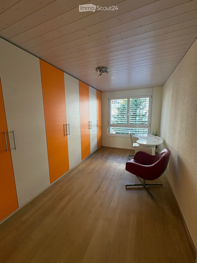 Bright room with wooden flooring, large window, modern orange and white cabinetry, and a small seating area with a chair and table.