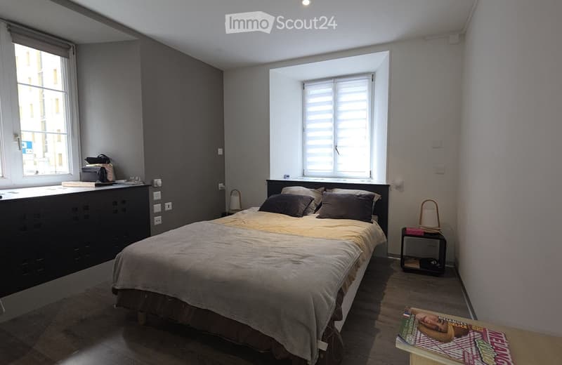 A modern bedroom with a large bed, nightstand, and a small table with a book on it. The room features wooden flooring, large windows with blinds, and a dark-colored wardrobe.