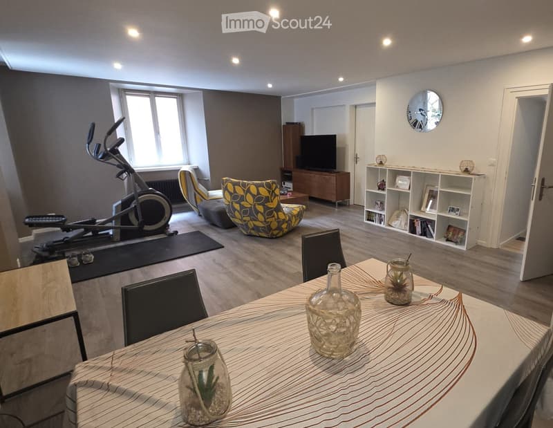 A modern living room with a large window, a dining table with chairs, a sofa, an armchair, a TV, a shelf, and an exercise bike in the corner.