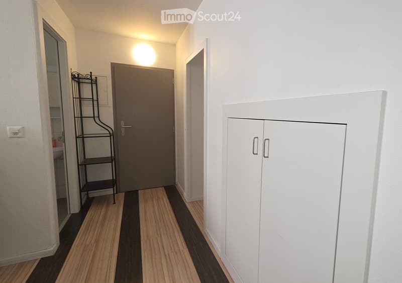 Empty hallway with wooden flooring, white walls, a closed door, a small black shelving unit, and a white cabinet with double doors.
