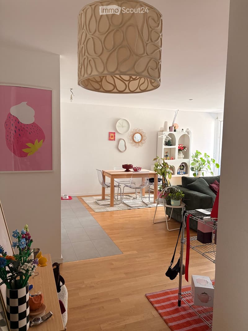 A modern living room with wooden flooring, a large pendant light, a green sofa, a wooden dining table with chairs, and various decorative items including a flower vase, a clock, and a shelf with plants.