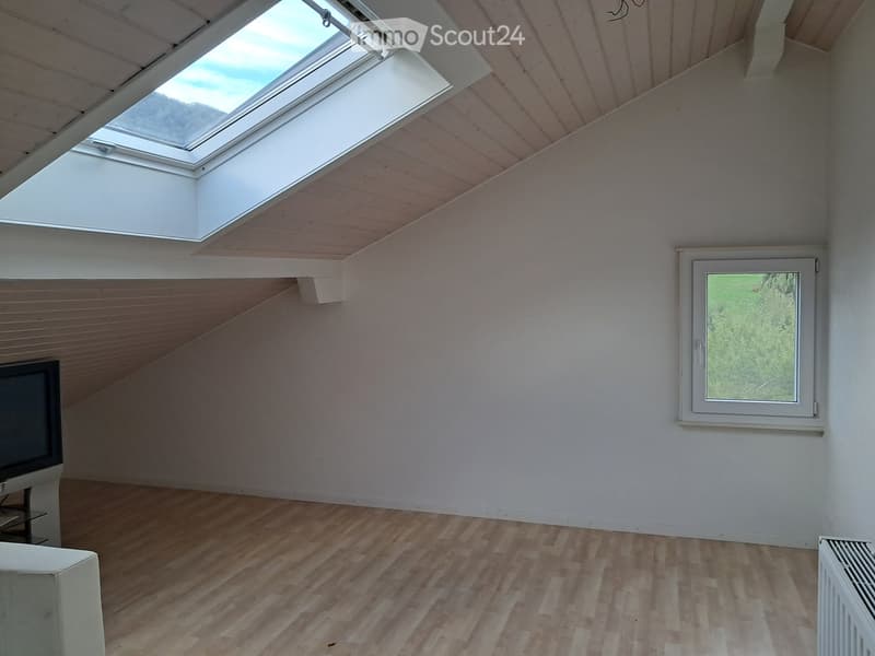 Empty attic room with wooden flooring, white walls, skylight windows, and a single window with a view of greenery.