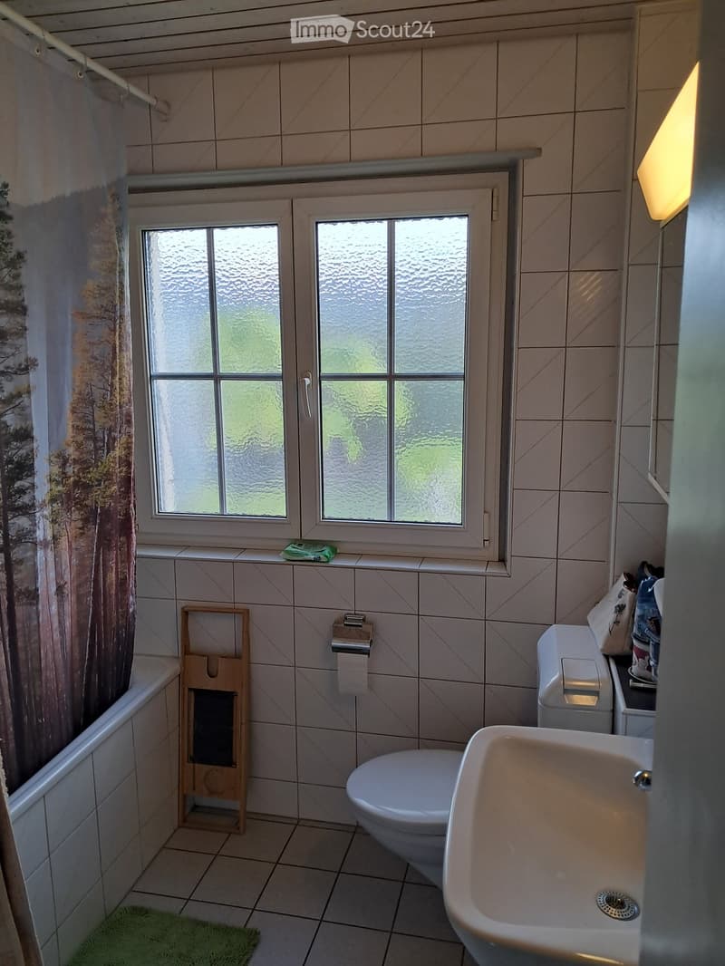 Bathroom with tiled walls and floor, featuring a bathtub, toilet, sink, and a window with frosted glass. A towel rack and a small green rug are also visible.
